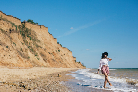 Happy young boho woman walking in sea waves in sunny warm day at tropical island and blue sky. Space for text. Stylish hipster girl in hat  relaxing on beach and smiling. Summer vacationの写真素材