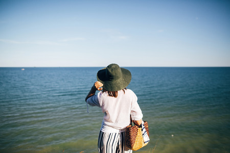 Stylish boho girl in hat looking at sea in sunny evening light from sandy cliff, back view. Happy young hipster woman relaxing on tropical island beach. Summer vacation. Space for textの写真素材