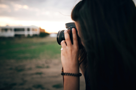 Stylish hipster girl taking photos on beach at sunset. Summer vacation. Portrait of  happy boho woman holding photo camera and making photos in evening time at  sea cliffの写真素材