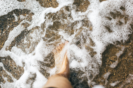 Feet standing in waves foam on sand, top view.  Man foot in warm water waves, relaxing on beach at sea. Summer vacation. Copy spaceの写真素材