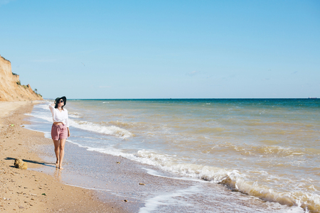 Stylish hipster girl in hat walking on beach and smiling. Summer vacation. Happy young boho woman relaxing and enjoying sunny warm day at tropical island and blue sky. Space for textの写真素材