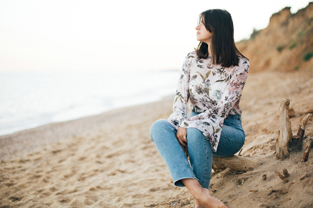 Stylish hipster girl sitting on beach at sea. Happy fashionable boho woman relaxing at sandy cliff on tropical island and looking at water. Travel and summer vacation. Space for textの写真素材