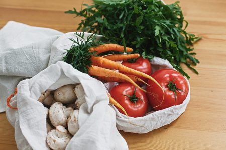 Fresh vegetables in eco cotton bags on wooden table in kitchen. Carrots,tomatoes, arugula, mushrooms from market in canvas reusable bags. Zero waste grocery shopping concept. ban plasticの写真素材