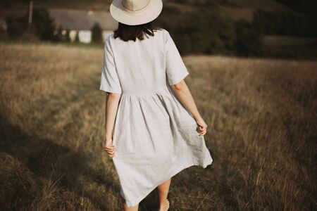 Stylish girl in linen dress and hat walking barefoot among herbs and wildflowers in sunny field in mountains. Boho woman relaxing in countryside, simple rustic life. Atmospheric imageの写真素材