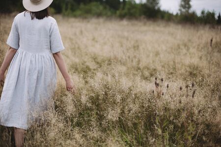 Stylish girl in linen dress and hat walking among herbs and wildflowers in field. Boho woman relaxing in countryside, simple slow life style. Space for text. Atmospheric imageの写真素材