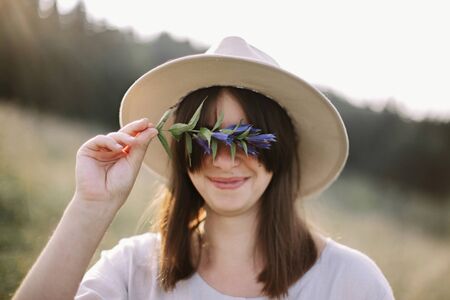 Stylish girl in rustic dress holding wildflower in hand, standing in sunny meadow in mountains. Boho...