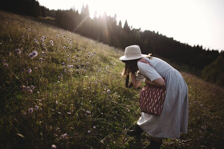 Stylish girl in rustic dress and hat taking photo of wildflowers  in sunny meadow in mountains. Boho woman relaxing in countryside at sunset, simple life. Atmospheric imageの写真素材