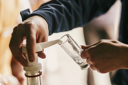 Waiter pouring vodka in glass on table at wedding reception. Guests hands holding alcohol drink or cocktail from big jar at alcohol bar at wedding or christmas feast. Luxury cateringの写真素材