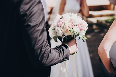 Stylish groom in black suit holding modern wedding bouquet of tender pink roses and peonies for bride in sunny morning. Wedding day preparationの写真素材
