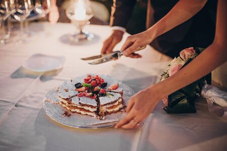 Stylish happy bride and groom cutting together wedding cake with fruits at wedding reception outdoors in the evening. Couple hands cutting cake in soft candle light in night.の写真素材