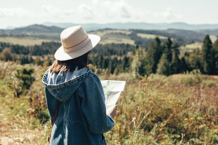 Stylish hipster girl holding map and traveling in sunny mountains. Woman in hat exploring map and hiking on top of mountain on summer vacation. Wanderlust and travel conceptの写真素材