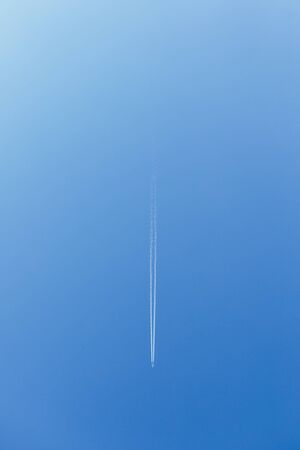 White trace of smoke from flying airplane. Vertical image. Plane flying in blue sky and living white...