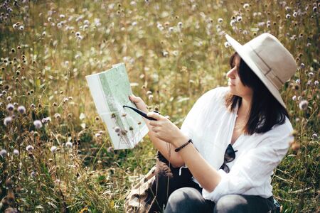 Hipster girl with wooden pipe looking at map and sitting in sunny  meadow, traveling in mountains. Stylish woman in hat hiking and exploring mountain. Wanderlust and travel conceptの写真素材