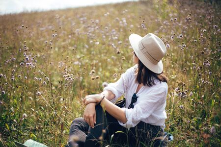 Hipster girl with backpack relaxing in wildflower meadow, traveling in sunny mountains. Stylish woman in hat enjoying hiking on top of mountain, sitting in herbs. Wanderlust and travelの写真素材