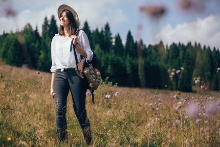 Wanderlust and travel concept. Hipster girl with backpack traveling in sunny mountains, walking in  wildflower meadow. Stylish happy woman in hat exploring and hiking on top of mountain.の写真素材