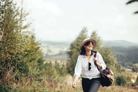 Happy hipster girl with backpack traveling on top of sunny mountain, walking on hills with woods. Stylish woman smiling and  enjoying hiking in mountains. Wanderlust and travel.の写真素材