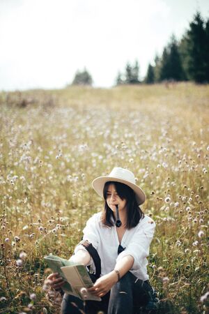 Hipster girl with wooden pipe looking at map and sitting in sunny  meadow, traveling in mountains. Stylish woman in hat hiking and exploring mountain. Wanderlust and travel conceptの写真素材
