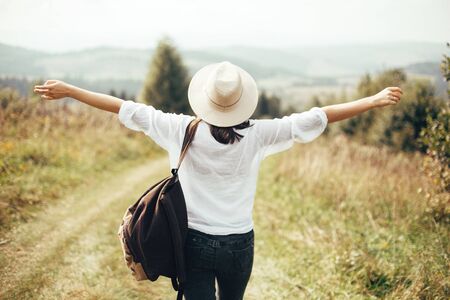 Hipster girl with backpack traveling on top of sunny mountain and raising hands, looking at hills and woods. Stylish woman enjoying hiking in mountains. Atmospheric momentの写真素材