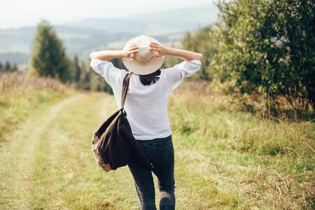 Hipster girl with backpack traveling on top of sunny mountain and looking at hills and woods. Stylish woman enjoying hiking in mountains. Wanderlust and travel. Atmospheric momentの写真素材
