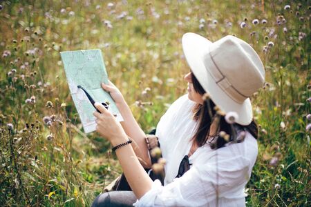Hipster girl with backpack looking at map and holding wooden pipe, sitting in wildflower meadow, traveling in sunny mountains. Stylish woman in hat hiking and exploring mountain.の写真素材