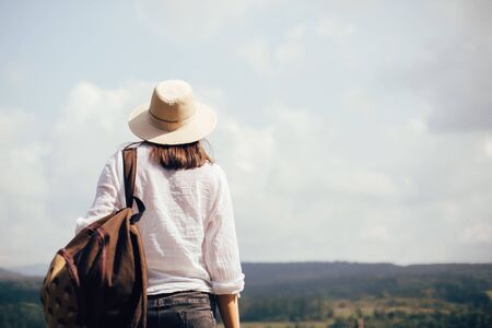 Hipster girl with backpack traveling and standing on top of sunny mountain and looking at sky and...