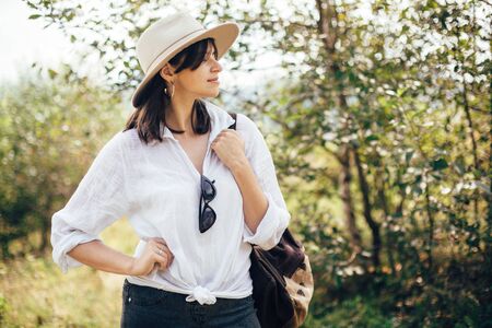 Hipster girl with backpack traveling on top of sunny mountain, relaxing in woods. Stylish woman smiling and enjoying hiking in mountains. Wanderlust and travel. Atmospheric momentの写真素材