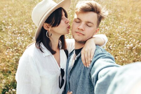 Happy hipster couple making selfie in sunny wildflower meadow, traveling in mountains. Stylish young family smiling and enjoying hiking on top of mountain, taking photo. Summer vacationの写真素材