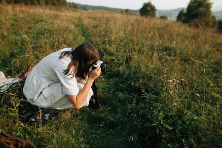 Stylish boho girl taking photo on film analog photo camera of grass and wildflowers in sunny meadow at sunset. Happy hipster woman enjoying traveling in evening mountainsの写真素材