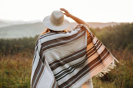 Stylish boho girl walking in sunny light at atmospheric sunset in meadow. Happy hipster woman in poncho and hat enjoying traveling in evening mountains. Space for textの写真素材