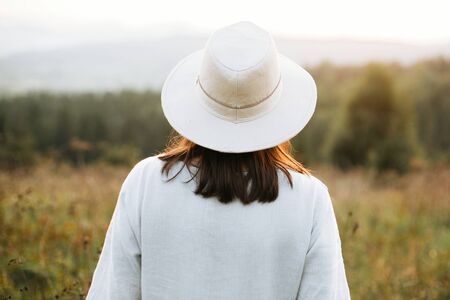 Stylish boho girl walking in sunny meadow at  atmospheric sunset. Happy hipster woman in linen...