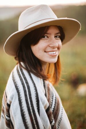 Stylish boho girl smiling  in sunny light at atmospheric sunset in meadow. Happy hipster woman in poncho and hat enjoying traveling in evening mountains. Space for textの写真素材
