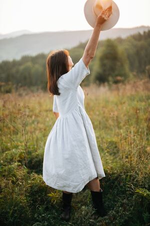 Stylish carefree boho girl throwing her hat in the sky in sunny light  at atmospheric sunset. Happy hipster woman in linen rustic dress enjoying traveling in evening  meadowの写真素材