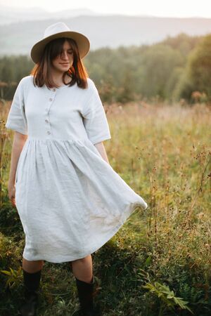 Stylish boho girl walking in sunny meadow at  atmospheric sunset. Happy hipster woman in linen rustic dress and hat enjoying traveling in evening mountains. Space for textの写真素材