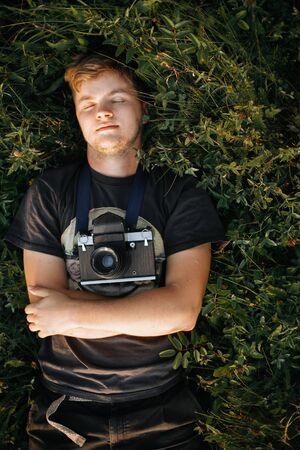 Stylish hipster man with film photo camera lying in green grass and relaxing in sunny meadow at...
