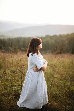 Stylish boho girl walking in grass and wildflowers in sunny meadow at  atmospheric sunset. Happy hipster woman in linen rustic dress enjoying traveling in evening mountains. Space for textの写真素材