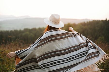 Stylish boho girl walking in sunny light at atmospheric sunset in meadow. Happy hipster woman in poncho and hat enjoying traveling in evening mountains. Space for textの写真素材