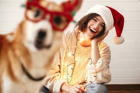 Stylish girl smiling in santa hat and christmas lights and looking at cute golden dog with funny emotions in festive reindeer glasses with antlers. Merry Christmas. Happy Holidaysの写真素材