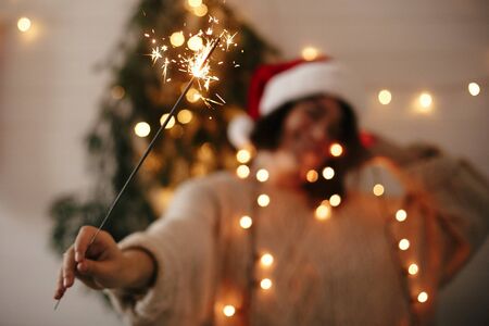 Happy New Year eve party atmosphere. Sparkler burning in hand of stylish girl in santa hat on background of modern christmas tree light in dark room. Woman with fireworks. Happy Holidaysの写真素材