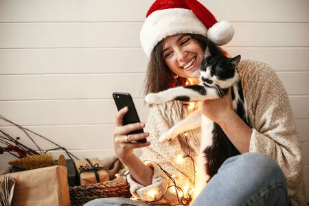 Stylish happy girl in santa hat taking selfie with cute cat in christmas lights on background of gifts. Young hipster woman in sweater hugging kitty with funny emotions at christmas treeの写真素材