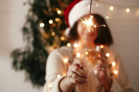 Happy New Year eve party atmosphere. Sparkler burning in hand of stylish girl in santa hat on...