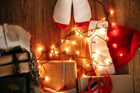 Top view of stylish festive socks, christmas gift boxes, santa hat, garland lights on wooden floor under christmas tree in dark room. Christmas eve atmosphere. Merry Christmas.の写真素材
