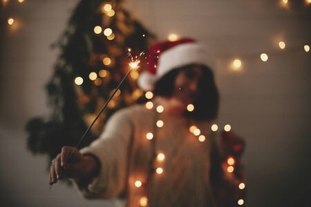 Big sparkler burning in hand of stylish hipster girl in santa hat on background of modern christmas tree light in dark room. Happy New Year eve atmosphere. Merry Christmas. Happy Holidaysの写真素材