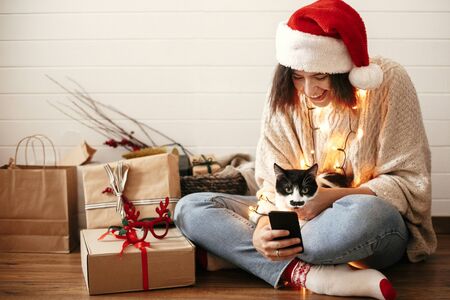 Stylish happy girl in santa hat with cute cat looking at phone screen on background of festive christmas lights and  presents. Young hipster woman hugging kitty with funny emotionsの写真素材