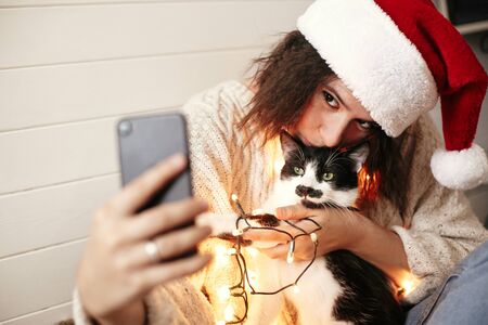 Stylish happy girl in santa hat taking selfie with cute cat in christmas lights on background of gifts. Young hipster woman in sweater hugging kitty with funny emotions at christmas treeの写真素材