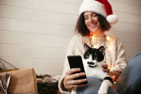 Stylish happy girl in santa hat with cute cat looking at phone screen on background of festive christmas lights and  presents. Young hipster woman hugging kitty with funny emotionsの写真素材