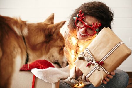 Stylish happy girl with christmas gift box in christmas lights and smiling to cute golden dog. Young woman in festive glasses with deer antlers  showing christmas present. Happy Holidaysの写真素材
