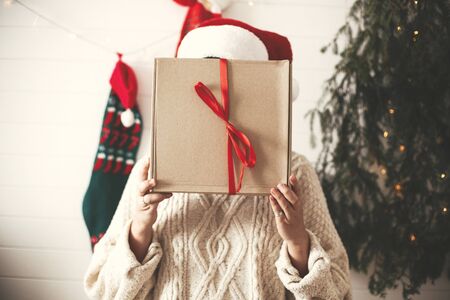 Stylish happy girl in santa hat holding christmas gift box in front of face on background of modern...
