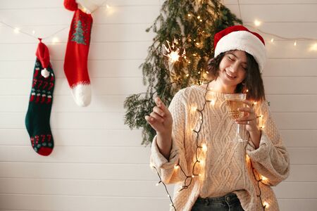 Stylish happy girl in santa hat holding sparkler and champagne glass, celebrating and toasting at christmas tree lights in festive room. Happy New Year eve party. Happy Holidaysの写真素材