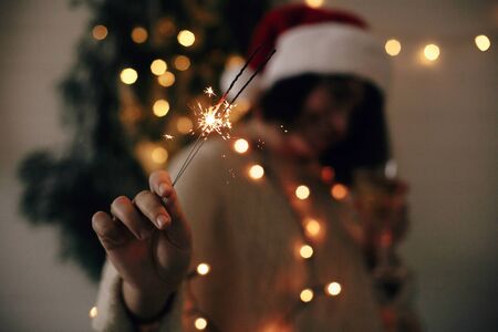 Big sparkler burning in hand of stylish girl in santa hat on background of modern christmas tree light in dark room. Woman with fireworks. Happy New Year eve atmosphere. Merry Christmasの写真素材
