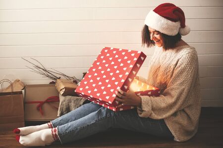 Stylish happy girl in santa hat and cozy sweater opening christmas gift box with magic light on...